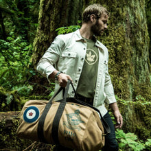 Load image into Gallery viewer, Man holding a tan RCAF duffel bag with a logo in a forest setting.