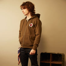Load image into Gallery viewer, Person wearing a brown hoodie with a red maple leaf design, standing against a beige wall.
